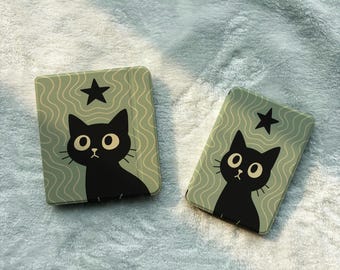 Pode incluir: Dois estojos retangulares com um fundo verde sálvia claro e um padrão de linhas onduladas. Cada estojo apresenta uma ilustração de um gato preto com olhos grandes e uma pequena estrela preta posicionada acima da cabeça do gato.