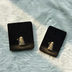 May include: Two black e-reader cases featuring a graphic illustration of a small, white, sheet-covered ghost holding a single yellow flower. The ghost is centered on a dark, shadowy background, creating a contrast between the light figure and the dark surroundings.