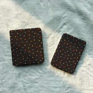 May include: Two rectangular electronic reading devices featuring dark brown protective covers. Both covers are decorated with a repeating pattern of small, multi-colored stars in shades of blue, orange, and red, distributed across the surface.