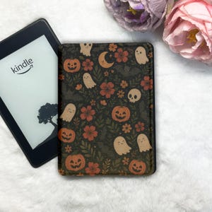 May include: A Kindle e-reader and a decorative cover with a Halloween design. The cover displays orange pumpkins, ghosts, flowers, and a crescent moon on a dark background. The Kindle screen shows a tree silhouette.
