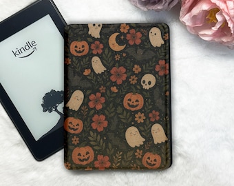 May include: A black e-reader case featuring a repeating pattern of orange jack-o'-lanterns, beige ghosts, skulls, crescent moons, and red flowers on a dark background, positioned next to a Kindle e-reader with a white screen.