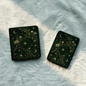 May include: Two rectangular e-reader cases featuring a dark forest green background with gold-toned botanical illustrations of swirling vines, white flowers, crescent moons, and radiating stars arranged in a symmetrical, intricate pattern.