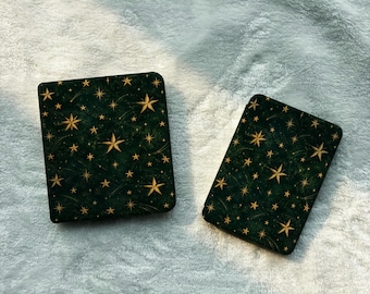 Può includere: Due custodie rettangolari con uno sfondo verde bosco scuro e un motivo ripetuto di stelle dorate e sottili linee radianti. Le custodie presentano angoli arrotondati e una finitura opaca liscia, con un design ispirato al cielo.