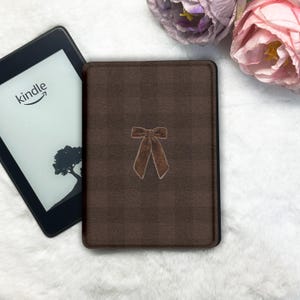 Puede incluir: Una funda marrón para Kindle con un diseño de lazo. La funda es rectangular y tiene una apariencia texturizada. Junto a la funda hay un lector de libros electrónicos Kindle con el logotipo de Kindle y una silueta de árbol en la pantalla.