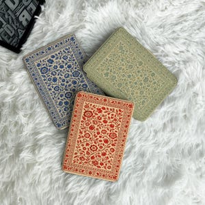 Könnte beinhalten: Drei rechteckige dekorative Schachteln mit floralen Mustern. Eine Schachtel ist blau mit weißen Blumen, eine andere grün mit blauen Blumen und die dritte rot mit roten Blumen. Die Schachteln sind auf einer weißen, flauschigen Oberfläche angeordnet.