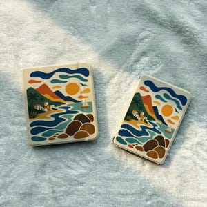 May include: Two square notebooks with a cream-colored cover featuring a colorful coastal landscape illustration. The design includes a bright yellow sun, blue ocean waves, brown rocky formations, green palm trees, and small white buildings.