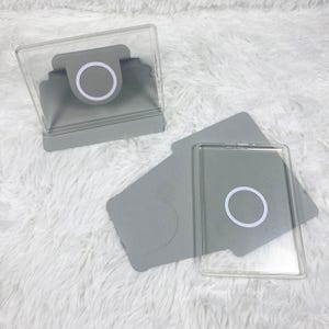 May include: Two grey protective cases for mobile devices, each featuring a clear rectangular frame and a circular white magnetic ring centred on the back. One case is positioned upright as a stand, while the other lies flat on its side.