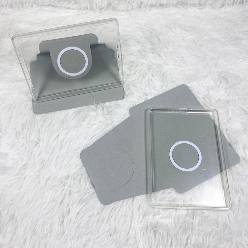 May include: Two grey protective cases for mobile devices, each featuring a clear rectangular frame and a circular white magnetic ring centred on the back. One case is positioned upright as a stand, while the other lies flat on its side.