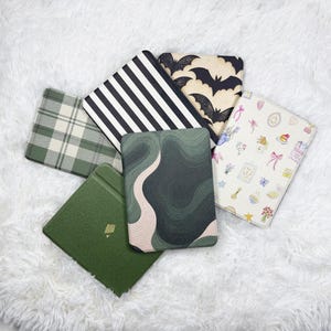 May include: A collection of colorful e-reader covers. Designs include a green plaid, black and white stripes, bats, a green and beige abstract pattern, and a cream-colored cover with floral and dessert illustrations.