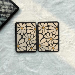 May include: A rectangular e-reader case featuring a repeating pattern of large, cream-coloured daisy flowers with yellow centres against a black background. The case is shown in an open, flat position, displaying the interior design on both panels.
