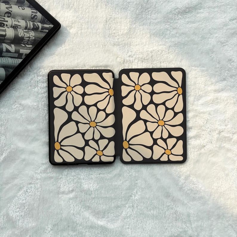 May include: A rectangular e-reader case featuring a repeating pattern of large, cream-coloured daisy flowers with yellow centres against a black background. The case is shown in an open, flat position, displaying the interior design on both panels.