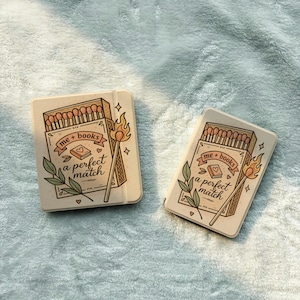May include: Two rectangular cases with a light beige finish, each featuring a printed illustration of a matchbox, a lit match, and green leaves. The text reads me + books a perfect match, accompanied by a small book icon and decorative hearts.