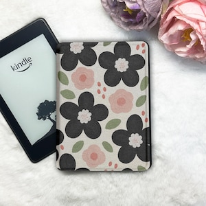 May include: Kindle e-reader next to a floral patterned cover. The cover features black and pink flowers, green leaves, and red dots on a cream background. The Kindle screen displays the word "kindle" and a tree silhouette.