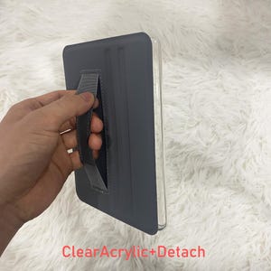 May include: A grey tablet case featuring a clear acrylic back and a detachable hand strap. The case has a smooth, matte finish on the front cover and includes the text ClearAcrylic+Detach printed in red at the bottom of the image.