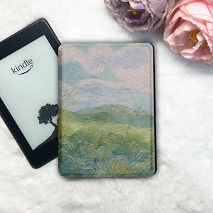 May include: A black Kindle e-reader with a screen displaying a silhouette of a tree and the word kindle, positioned next to a protective cover featuring a textured, impressionistic landscape painting of green fields and a pale sky.