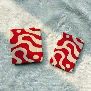 May include: Two rectangular electronic reading devices featuring a vibrant red and cream wavy, abstract pattern across their exterior shells. The devices are positioned side-by-side, displaying a consistent fluid design with circular accents.