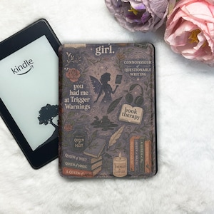 Op de afbeelding: Een Kindle e-reader naast een decoratieve hoes met een grillig ontwerp. De hoes toont een feeënsilhouet, rozen, boeken en tekst met de woorden "girl", "Trigger Warnings" en "book therapy".