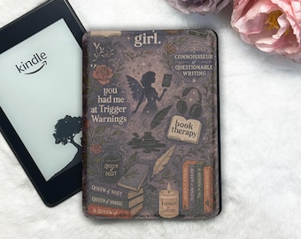 May include: A Kindle e-reader next to a decorative case with a floral and fairy design. The case features text including "girl," "you had me at Trigger Warnings," and "book therapy." The case has a dark purple background with illustrations.