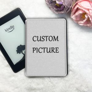 May include: A black Kindle e-reader with a tree graphic on the screen next to a light gray protective case featuring the black text CUSTOM PICTURE printed in the center. The case has a smooth, matte finish and rounded corners for the device.