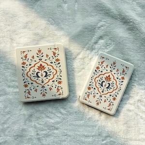 May include: Two rectangular notebooks with off-white covers featuring a symmetrical floral pattern in burnt orange and dark blue. The design includes intricate vines, leaves, and central petal motifs printed on the front of each book.