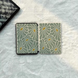 May include: A rectangular e-reader case featuring a repeating pattern of sage green wavy-petaled flowers with mustard yellow centres. The case is shown in an open position, displaying the interior design on both panels against a white textured surface.