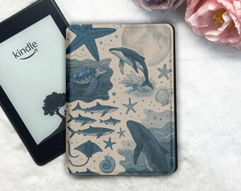 Puede incluir: Un lector de libros electrónicos Kindle con una funda azul y beige con ilustraciones de vida marina, incluyendo ballenas, tiburones y estrellas de mar. La pantalla del Kindle muestra el logotipo de Kindle y una silueta de árbol.