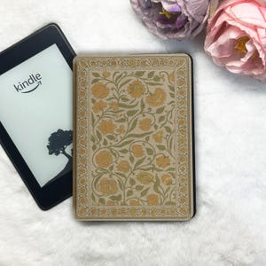 Może przedstawiać: Czytnik e-booków Kindle z dekoracyjnym etui z kwiatowym wzorem w odcieniach złota i zieleni. Kindle jest czarny, a etui ma szczegółowy wzór. Słowo "kindle" jest widoczne na ekranie.