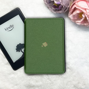 May include: A Kindle e-reader with the Amazon logo and a tree graphic is next to a green textured cover. The cover has a small decorative design. The background is a white, fluffy surface with pink and purple flowers.