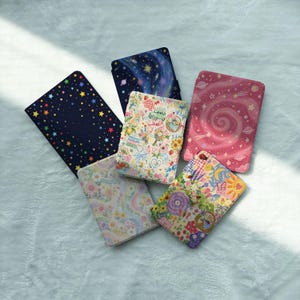 May include: Five rectangular e-reader cases featuring diverse designs: one with multicolored stars on black, one with a blue galaxy print, one with a pink spiral nebula, one with a pastel floral pattern, and one with a vibrant garden motif.