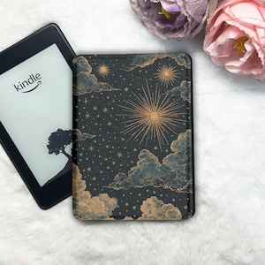 Könnte beinhalten: Ein Kindle E-Reader neben einer dekorativen Hülle. Die Hülle hat ein dunkelblaues Himmelsdesign mit goldenem Sonnenaufgang, Sternen und Wolken. Der Kindle-Bildschirm zeigt das Wort "Kindle" und eine Baumsilhouette.