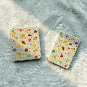 May include: Two rectangular cases with a cream-colored background featuring a black polka dot pattern and a variety of colorful fruit illustrations including pineapples, strawberries, bananas, cherries, blueberries, and slices of melon and papaya.