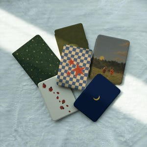 May include: A collection of six rectangular e-reader cases featuring various designs: a dark green starry pattern, a solid olive green, a blue and white checkerboard with red stars, a white case with ladybugs, a dark blue with a gold moon, and a landscape.