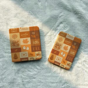 Op de afbeelding: Twee rechthoekige hoezen voor elektronische apparaten met een rasterpatroon in tinten oranje, bruin en crème. Het ontwerp bevat iconen van bladeren, geesten, pompoenen en enveloppen, met de tekst VIBES ONLY, FALL en sweater weather.
