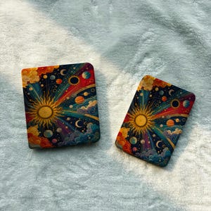 May include: Two rectangular cases featuring a vibrant celestial design. The artwork displays a central golden sun with radiating beams, surrounded by colorful planets, stars, and swirling clouds against a deep blue cosmic background.