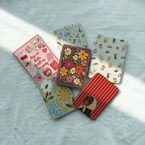 May include: Five rectangular e-reader cases with various designs: one with pink cherry and book motifs and text reading 'buy me books & tell me I'm pretty', one with blue stripes and fruit, one with coffee icons, one with a floral pattern, and one with red stripes and a dog.
