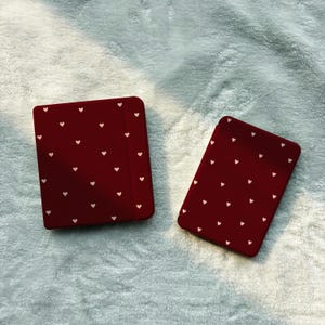 Könnte beinhalten: Zwei rechteckige elektronische Lesegeräte mit dunkelroten Hüllen, die mit einem sich wiederholenden Muster aus kleinen, hellrosa Herzen verziert sind. Die Geräte haben abgerundete Ecken und eine glatte, matte Oberfläche, wobei eines etwas größer ist.