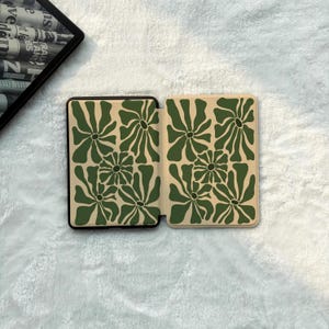 May include: A rectangular e-reader case featuring a repeating pattern of dark green abstract flower shapes on a light beige background. The case is shown in an open position, displaying the interior design on both the left and right panels.
