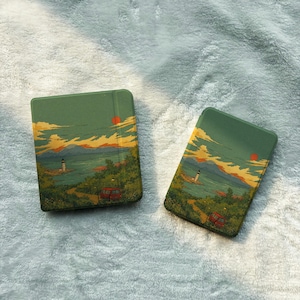 May include: Two rectangular cases featuring a printed landscape illustration of a coastal scene with a lighthouse, a red vehicle, and a bright orange sun against a green sky. The artwork depicts rolling hills, trees, and a calm body of water.