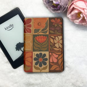 Puede incluir: Un lector de libros electrónicos Kindle con una funda estampada con flores. La funda presenta una cuadrícula de nueve cuadrados, cada uno con un diseño de flor diferente en tonos naranja, marrón y beige. La pantalla del Kindle muestra el logotipo de Amazon y una silueta de árbol.
