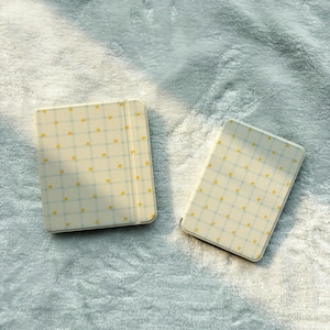 May include: Two square electronic reading devices featuring a light cream-colored exterior with a soft blue grid pattern and small yellow star icons scattered across the surface. Both devices have a smooth, matte finish and rounded corners.