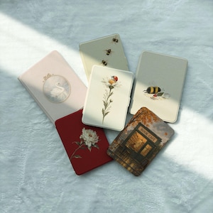 May include: Five rectangular e-reader cases featuring various designs: a pale pink case with a swan, a cream case with bees, a cream case with a daisy and ladybug, a cream case with a bee and tea, a red case with a peony, and a book cafe scene.