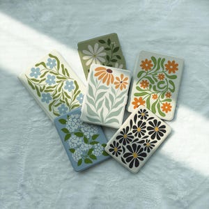 May include: Five rectangular e-reader cases featuring various floral patterns. One case has light blue flowers, one has white daisies on green, one has orange flowers, one has white blossoms on blue, and one has black daisies with yellow centres.