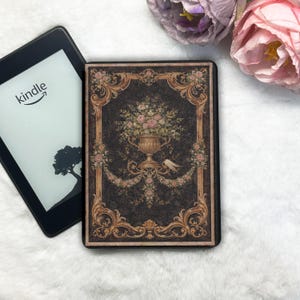 May include: Kindle e-reader with a decorative cover featuring a floral arrangement in a gold urn. The cover has a dark background with ornate gold trim and floral accents. The Kindle screen displays the Kindle logo and a tree silhouette.