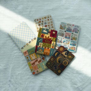 May include: Five rectangular notebooks with varied cover designs: one with a light blue checkerboard and cat faces, one with small kitchen icons, one with a patchwork of nature and building motifs, one with a grid of animals and fruit, and one with celestial patterns.