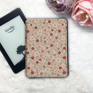 May include: A Kindle e-reader with a floral patterned cover featuring red and peach flowers on a beige background. The Kindle is black and displays the word "kindle" and a tree silhouette. The cover is open and next to the Kindle.