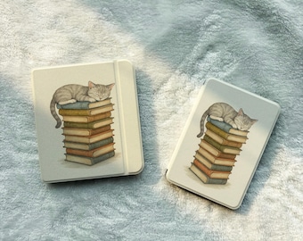 Pode incluir: Duas capas para leitor de e-books em tom off-white, cada uma com uma ilustração impressa de um gato malhado cinzento a dormir sobre uma pilha vertical de livros coloridos. As capas têm um design retangular liso com cantos arredondados.