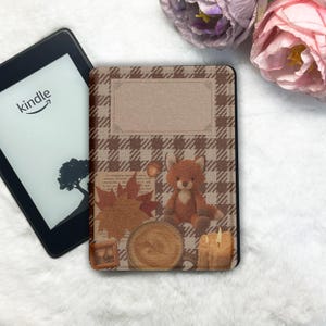 May include: A Kindle e-reader next to a decorative case with a brown plaid pattern. The case features a fox plush, autumn leaves, a coffee cup, and a candle. The Kindle screen displays the Amazon logo and a tree silhouette.