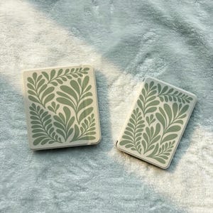 May include: Two rectangular cases with a cream-colored background and a green leaf design. The cases are lying on a soft, light blue surface. The leaf design is a repeating pattern of stylized leaves and branches.