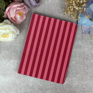 May include: A rectangular, red and pink striped case, likely for an e-reader or tablet, is displayed on a gray surface. The case has a vertical stripe pattern in two shades of red. The image is styled with floral accents.