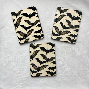 May include: Three rectangular tablet cases featuring a repeating pattern of black bats on a cream-colored background. Each case displays a variety of bat silhouettes, some with intricate wing details, arranged in a dense, overlapping design across the surface.